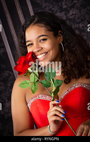 Uno splendido, biracial high school girl senior indossando red party dress il trattenimento di una rosa rossa Foto Stock