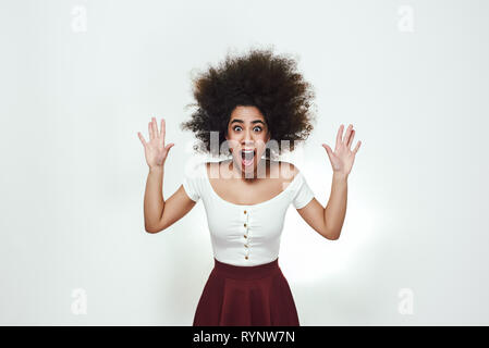 Effetto Wow! Sorpresi e totalmente sconvolta afro american donna è il sollevamento in alto le mani mentre in piedi in studio contro uno sfondo grigio con la bocca aperta. Sconvolgente notizia Foto Stock