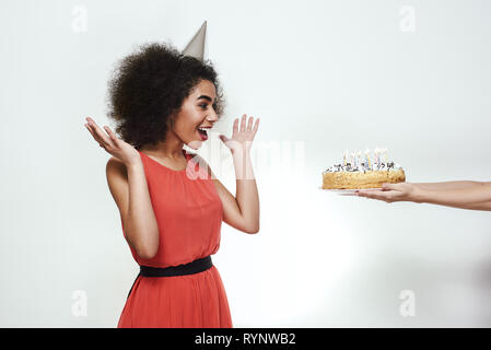 Sorpresa per voi! Entusiasti giovani afro american donna in party hat alzando le mani e guardando alla torta di compleanno con candele Foto Stock