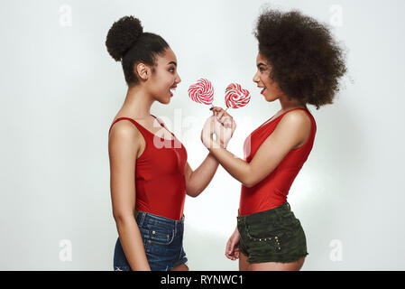 Provare il mio lecca-lecca! Due giovani allegri afro american donna in abiti estivi sono holding lecca-lecca e guardando gli uni con gli altri mentre in piedi contro uno sfondo grigio. Concetto di divertimento. La bellezza delle donne Foto Stock