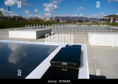Telefono cellulare la carica in remoto su un banco solare con fontane e cielo blu in background. Foto Stock