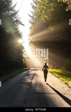 Giovane donna jogging lungo una strada, la luce del sole splende attraverso gli alberi, Oregon Coast Highway, Oregon, Stati Uniti d'America Foto Stock