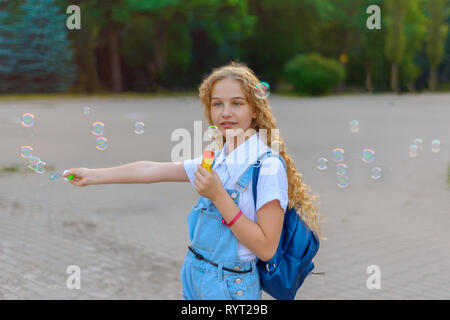 Ragazza bionda con capelli ricci in tuta in jeans sorridenti soffia bolle di sapone nel parco Foto Stock