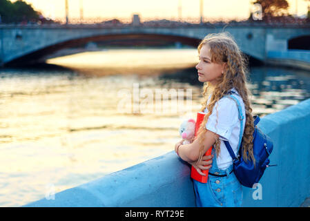 La ragazza è in piedi vicino al fiume in città al tramonto. Un adolescente con uno zaino è in possesso di un orso giocattolo e una cartella rossa Foto Stock