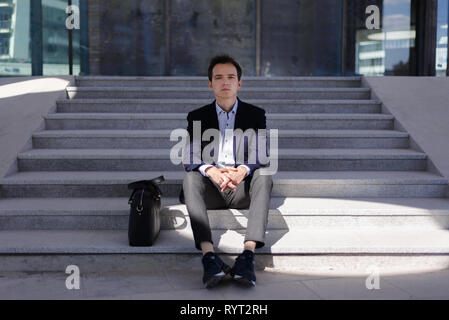 Ragazzo giovane imprenditore triste e pensieroso. l uomo in giacca blu e camicia è seduto sui gradini accanto a valigetta Foto Stock