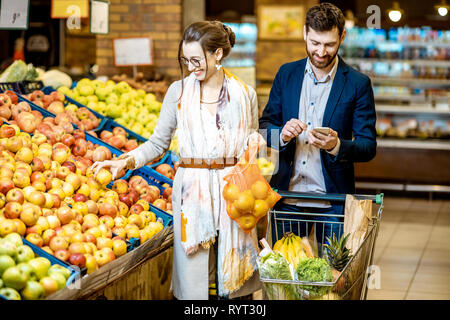 Giovane e felice coppia acquistare frutta e verdura fresca in piedi insieme con il carrello del supermercato Foto Stock