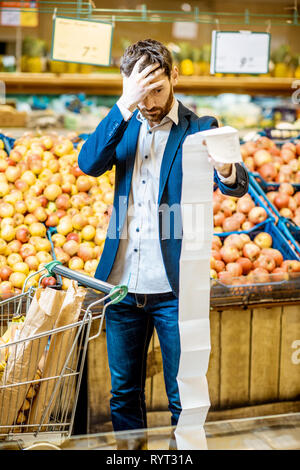 Ritratto di un uomo elegante con scioccato emozioni tenendo molto lunga lista di shopping mentre l'acquisto di alimenti nei supermercati Foto Stock