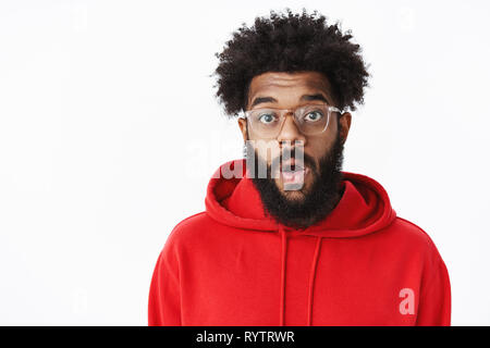 Close-up shot di sorpreso stupiti e senza parole americano africano maschio barbuto con acconciatura afro in bicchieri e felpa con cappuccio rosso caduta dalla ganascia Foto Stock