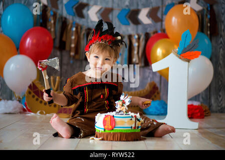 Foto riprese smash la torta per 1 anno in stile di capo indiano. Little Boy emotivo con un tomahawk in costume di Apache e in piuma copricapo Foto Stock