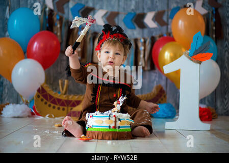 Foto riprese smash la torta per 1 anno in stile di capo indiano. Little Boy emotivo con un tomahawk in costume di Apache e in piuma copricapo Foto Stock