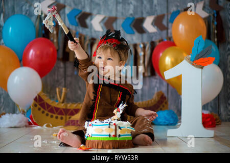 Foto riprese smash la torta per 1 anno in stile di capo indiano. Little Boy emotivo con un tomahawk in costume di Apache e in piuma copricapo Foto Stock