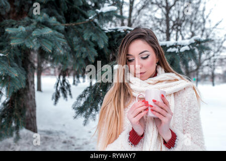 Bellissima ragazza con i capelli lunghi in inverno nel parco cittadino, tenendo la tazza di caffè caldo il tè con la sua mano, il riscaldamento se stessa. Sogni fantasticare, spazio libero per Foto Stock