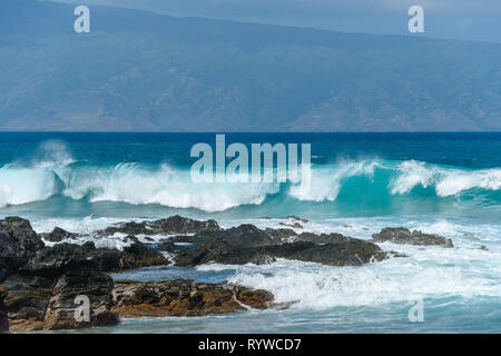 Le onde che si infrangono - grande rottura onde blu precipitando verso il nero costa rocciosa con un isola tropicale in background. Maui, Hawaii, Stati Uniti d'America. Foto Stock