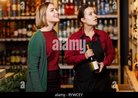 Foto di due giovani donne su sfondo sfocato di scaffali con bottiglie di vino Foto Stock