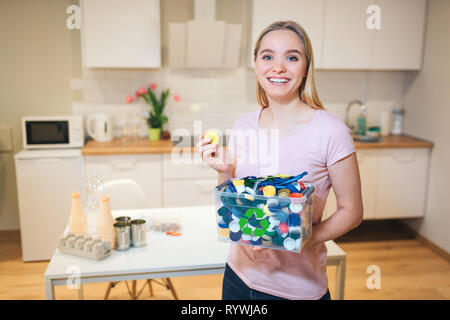 Il riciclaggio. Giovane donna sorridente holding coperchi in plastica nel contenitore con riciclo verde icona sullo sfondo di cucina Foto Stock