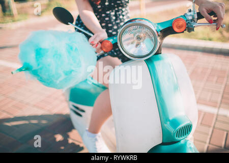 Giovani felici hipster donna mangiare zuccherato caramella di cotone. Modello femminile in sella a un scooter blu una strada di città Foto Stock