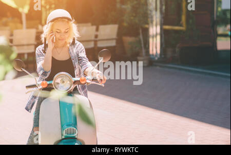 Bella giovane donna vestiti casual seduto su uno scooter e parlando al telefono. Ragazza seduta sulla moto e utilizzando cellulari smartphone Foto Stock