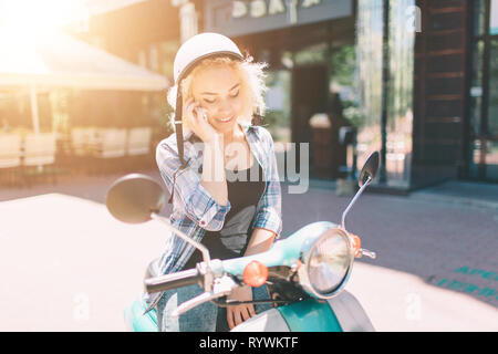 Bella giovane donna vestiti casual seduto su uno scooter e parlando al telefono. Ragazza seduta sulla moto e utilizzando cellulari smartphone Foto Stock