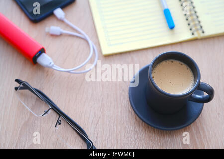 Lo smartphone è in carica dal poverbank. Tazza di caffè, agenda, matita e bicchieri in tavola close-up Foto Stock
