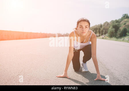 Donna giovane atleta alla posizione di partenza pronta per iniziare una corsa. Velocista femmina pronto per l'esercizio sportivo su autodromo con sun flare. Foto Stock