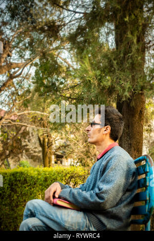 Immagine esterna di asiatici, indiani serena giovane imprenditore adulto seduto su una panchina nel parco, tenendo business planner e pensare guardando lontano a vista Foto Stock