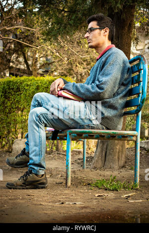 Immagine esterna di asiatici, indiani serena giovane imprenditore adulto seduto su una panchina nel parco, tenendo business planner e pensare guardando lontano a vista Foto Stock