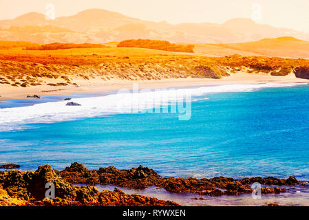 Fumoso estate spiaggia e le montagne al di là sotto oro brillante cielo nebuloso. Pomeriggio di sole sulla spiaggia di sabbia dorata, oceano blu luminoso con il bianco onde. Foto Stock