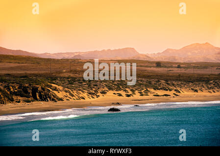 Opaco giallo arancione pomeriggio cielo sopra le montagne nebuloso e la spiaggia di sabbia fine e dorata con blu oceano. Onde che si infrangono sulla spiaggia. Foto Stock