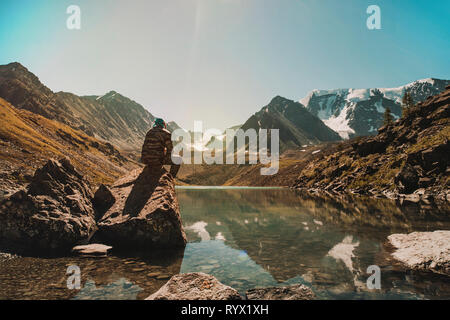 Viaggiatore giovane uomo si siede su una pietra e si ammira il bellissimo lago di montagna nelle montagne di Altai. La Siberia in Russia. Un uomo in camuffamento unifor militare Foto Stock