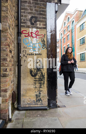 Una giovane donna che indossa una felpa con cappuccio passato a piedi un pezzo di Rude Kids graffiti su una strada di Londra, Regno Unito Foto Stock