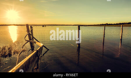 Palo di legno all'interno del lago con i riflessi del sole nell'acqua al tramonto e il cielo arancione con le nuvole in background Foto Stock