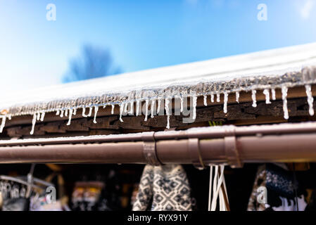 Ghiaccioli pendenti dal tetto di un edificio in legno su un inverno gelido giorno, un sacco di neve sul tetto, un visibile grondaia in plastica e cielo blu. Foto Stock