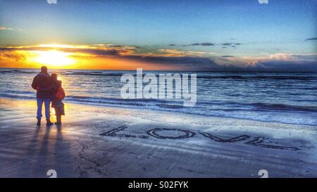 Un paio di stand a riva a guardare il tramonto la parola amore inscritta nella sabbia accanto a loro Foto Stock
