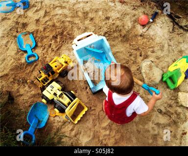 Il Toddler boy gioca con una pala e carrelli in una sandbox di grandi dimensioni. Foto Stock