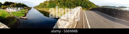 Panoramica del Clachan Bridge o un ponte sull'Atlantico a Seil isola vicino Easdale e Oban in Scozia che mostra la carreggiata del ponte e il vicino cottage bianco accanto al mare. Foto Stock