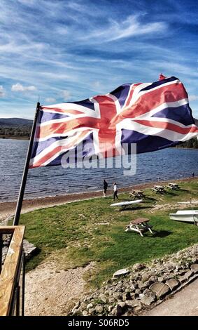 Union Jack Flag di Gran Bretagna e Irlanda del Nord battenti; sparato contro un scozzese loch lago. Formato verticale Foto Stock