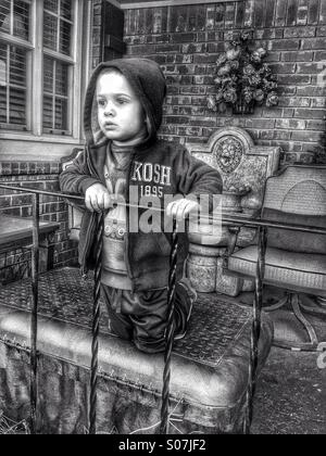 4 anno vecchio ragazzo appoggiata su un portico ringhiera e apparentemente pensando seriamente. HDR in bianco e nero Foto Stock