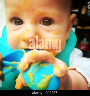 Baby boy quasi 6 mesi le frizioni cucchiaino per lo svezzamento come egli tenta di mangiare purea di zucca Foto Stock