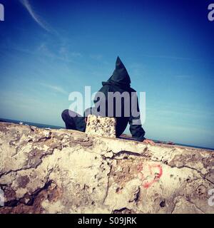 Uomo che indossa djellaba, seduti nella parete a Essaouira, UNESCO World Heritage Town, Marocco Foto Stock