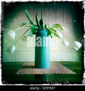 L'avvizzimento tulipani in un blu/verde vaso stagno contro un dipinto di bianco parete in legno su un tappeto di fiori su un tavolo in legno di quercia. Foto Stock