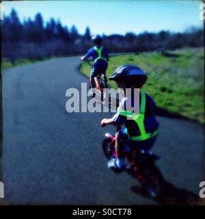 Due ragazzi in bicicletta sul percorso in un parco in una giornata di sole indossare giubbotti di sicurezza e caschi Foto Stock