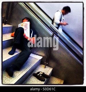 Un " commuter " guardando il suo smartphone passa da un uomo prendendo un pisolino in Raffles Place MRT Station nel quartiere centrale degli affari di Singapore Foto Stock