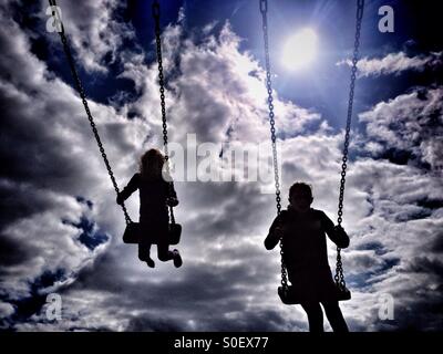 Due bambini su altalene in silhouette con il sole alle spalle Foto Stock