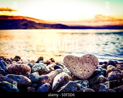 Amore di estate. Evidenziare la forma di cuore rock seduto sulla spiaggia in primo piano. Tramonto sul lago e le montagne sullo sfondo. Foto Stock