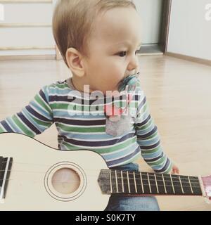 Baby boy a suonare la chitarra , guardando al lato, seduto su un pavimento di legno in una stanza vuota con un ciuccio in bocca Foto Stock