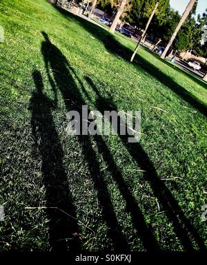 Lungo, Tall ombre sull'erba di un genitore tenendo le mani con due bambini Foto Stock