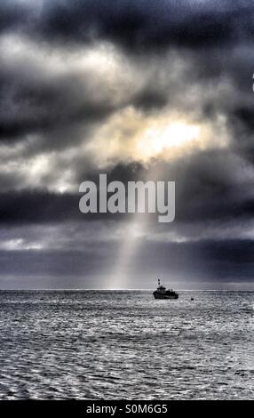 Un albero di luce del sole si rompe in un nero cielo tempestoso mentre una solitaria barca da pesca galleggianti sul mare sottostante. Foto Stock