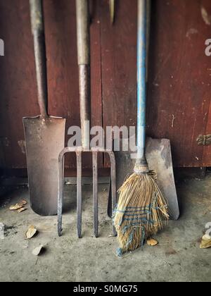 Scopa e arrugginiti attrezzi da giardino in appoggio contro un antico armadio in legno. Foto Stock