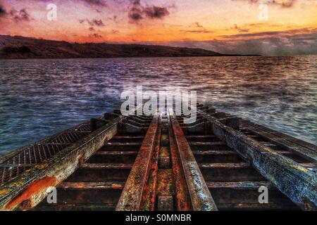 Un arrugginito scialuppa di salvataggio di uno scalo in uno stato di degrado scompare in un acciaio, grigio oceano a sunrise. Foto Stock