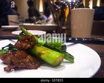 Vista ravvicinata del manzo e del gai-lan (broccoli cinesi) in un ristorante cinese su un piatto bianco, mentre altri commensali sono fuori fuoco Foto Stock